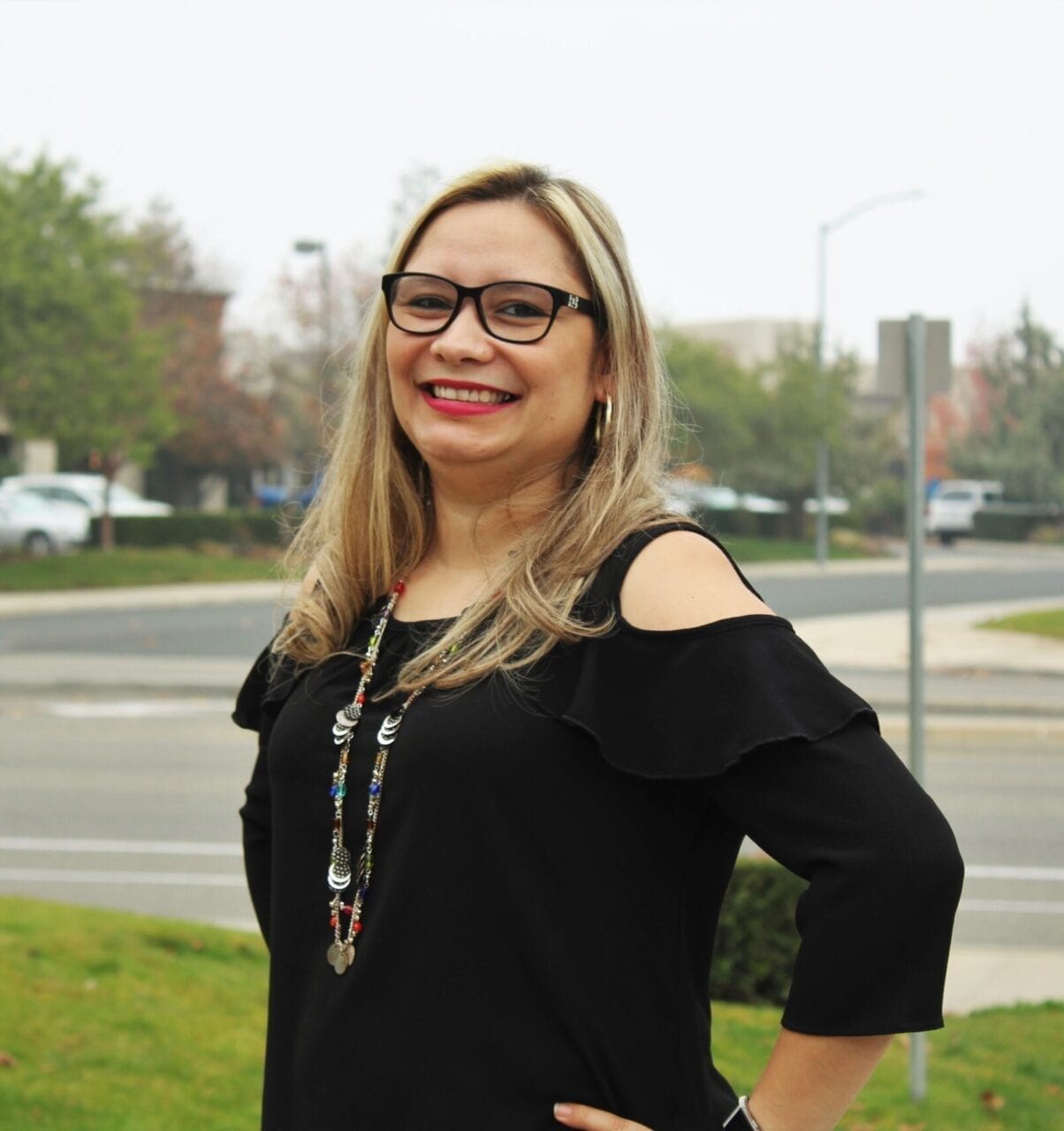 Smiling woman with glasses standing outdoors in a casual black outfit.